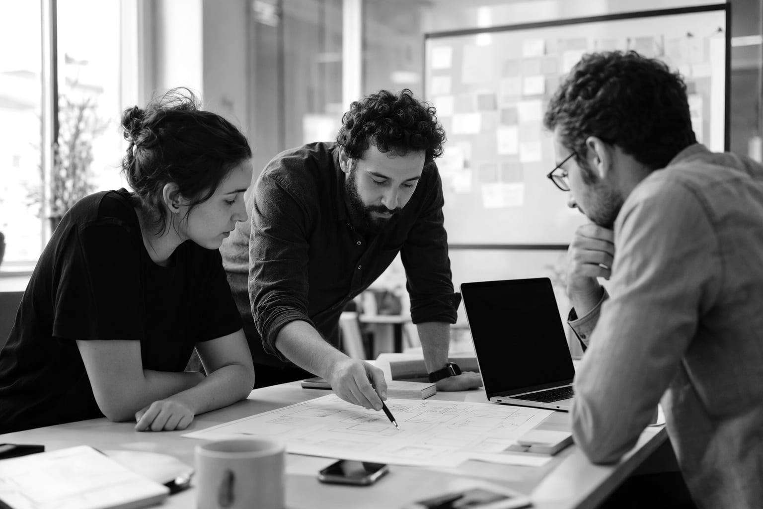 Three colleagues gathered around a desk, one pointing at a printed diagram while the others look on, a laptop and a pinboard of notes nearby