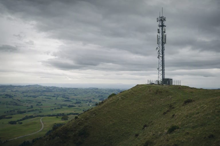 A cellular tower on a green hilltop under a moody grey sky, with farmland below