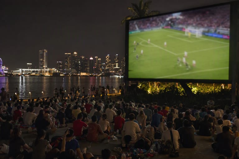 A crowd watches live football on a large outdoor screen beside a lit-up waterfront at night