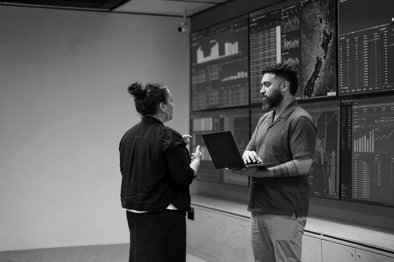 An operations room with a wall of monitors; two people stand talking and one holds a laptop